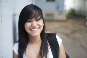 An image of a young college student smiling and leaning against a wall.
