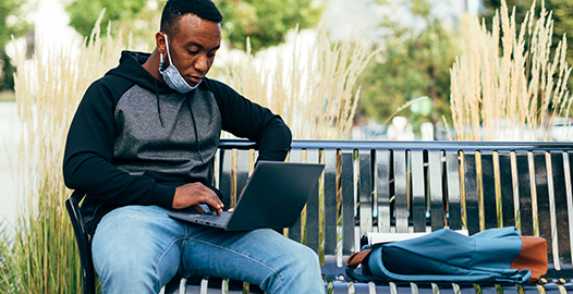An image of a young man sitting on a park bench and working on his laptop.