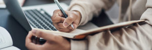 An image of a student taking notes on a pad while working on their laptop.