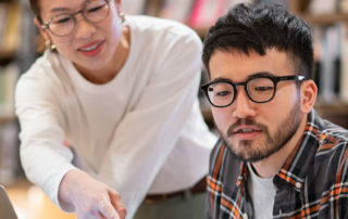 An image of a college professor helping a student at his laptop.