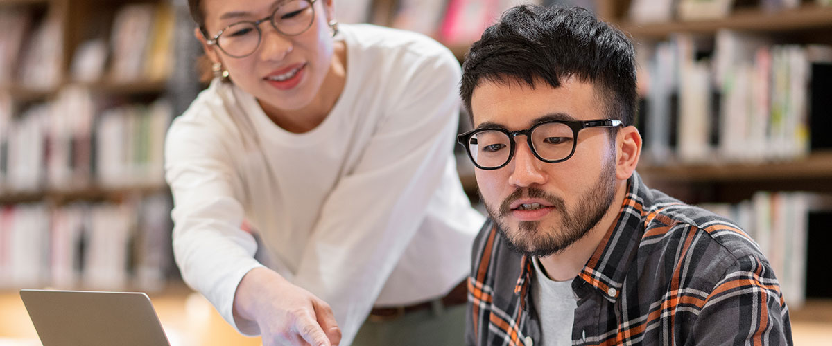 An image of a college professor helping a student at his laptop.